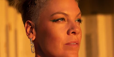 A person with short hair and green eyeliner looks into the distance, wearing a dangling earring, with warm sunlight illuminating their face.