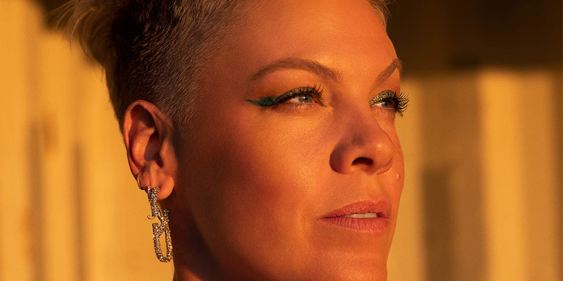 A person with short hair and green eyeliner looks into the distance, wearing a dangling earring, with warm sunlight illuminating their face.