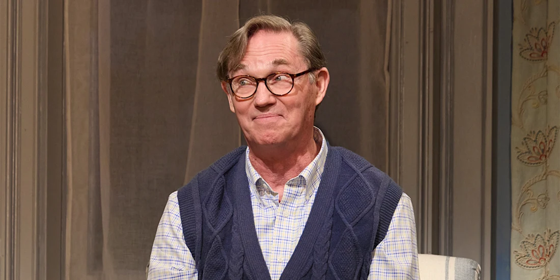 A man with glasses and a blue sweater vest sits indoors, smiling slightly with a neutral background behind him.