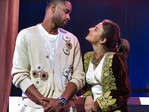 A man and a woman stand close together, looking at each other and smiling, both dressed in detailed, patterned clothing on a stage.