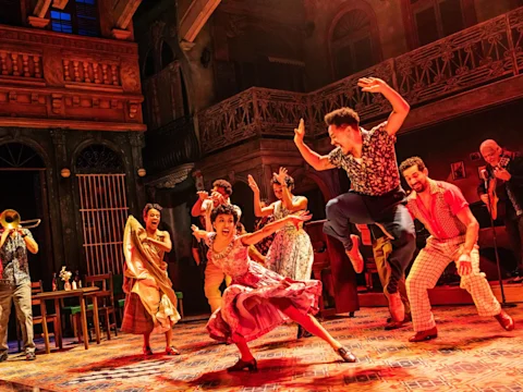 Actors in colorful costumes energetically dance and leap on stage, set against a vibrant, warmly lit backdrop with a balcony and musicians playing instruments.