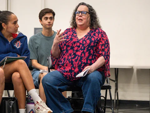 A woman in a red and blue patterned blouse speaks while seated among a group of people, holding notes in her hand.