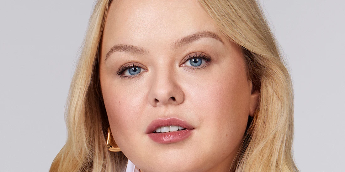 A woman with long blonde hair, light skin, and blue eyes looks directly at the camera. She is wearing a white top and gold hoop earrings against a plain background.