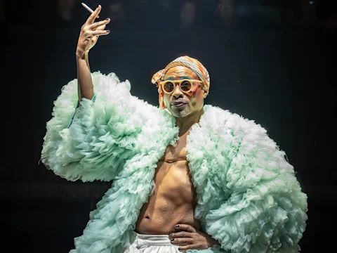 Person wearing a large, ruffled mint green robe, orange glasses, and a headscarf, holding a cigarette with an expressive hand gesture.