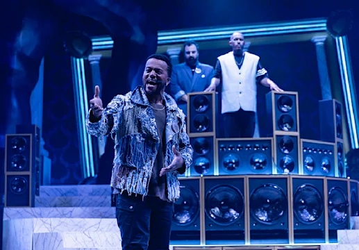 A man in a fringe jacket performs passionately on stage, with two men standing behind him among large speakers and columns under blue lighting.