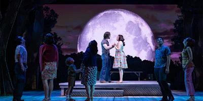 A group of people stand on a stage with a large, illuminated moon backdrop. Two people hold hands at center stage, while others watch.