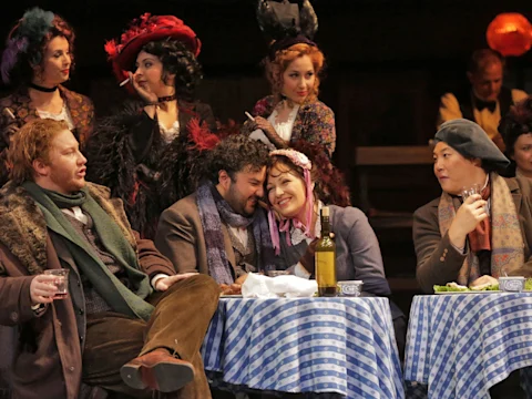 A group of people in period costumes sit around tables covered with blue-checkered cloths, engaged in lively conversation and interaction.