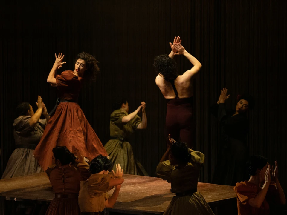 A group of people perform a dance routine on a wooden platform. Some wear flowing dresses while others wear dark clothing. They are captured in mid-movement with hands raised.