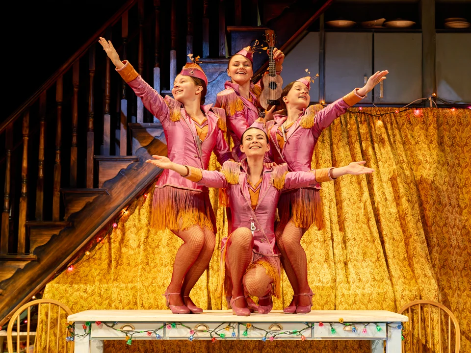 Four performers in uniform-style outfits pose with outstretched arms and smiles on stage, with one person holding a violin. Curtain backdrop and stage lights visible.