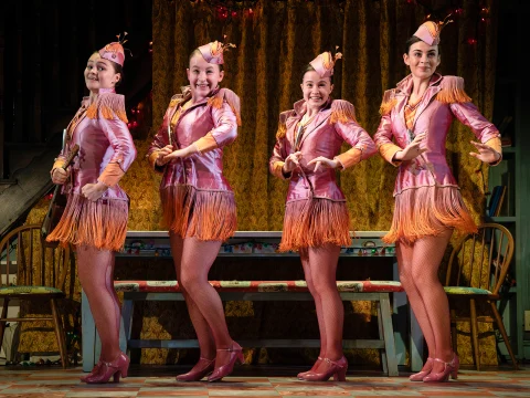 Four performers in matching pink and orange outfits strike a pose on stage, against a gold curtain backdrop.