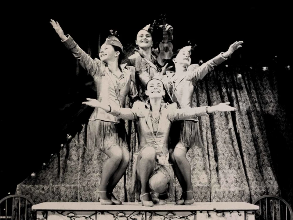 Four performers in uniform-style outfits pose with outstretched arms and smiles on stage, with one person holding a violin. Curtain backdrop and stage lights visible.