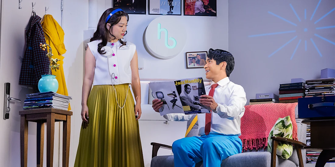 A woman stands and looks at a seated man who is reading a magazine in a brightly lit, modern living room with books and decor.