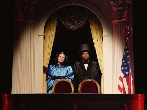 Two people dressed in historical costumes stand in a theater box with gold curtains and an American flag visible to the right.