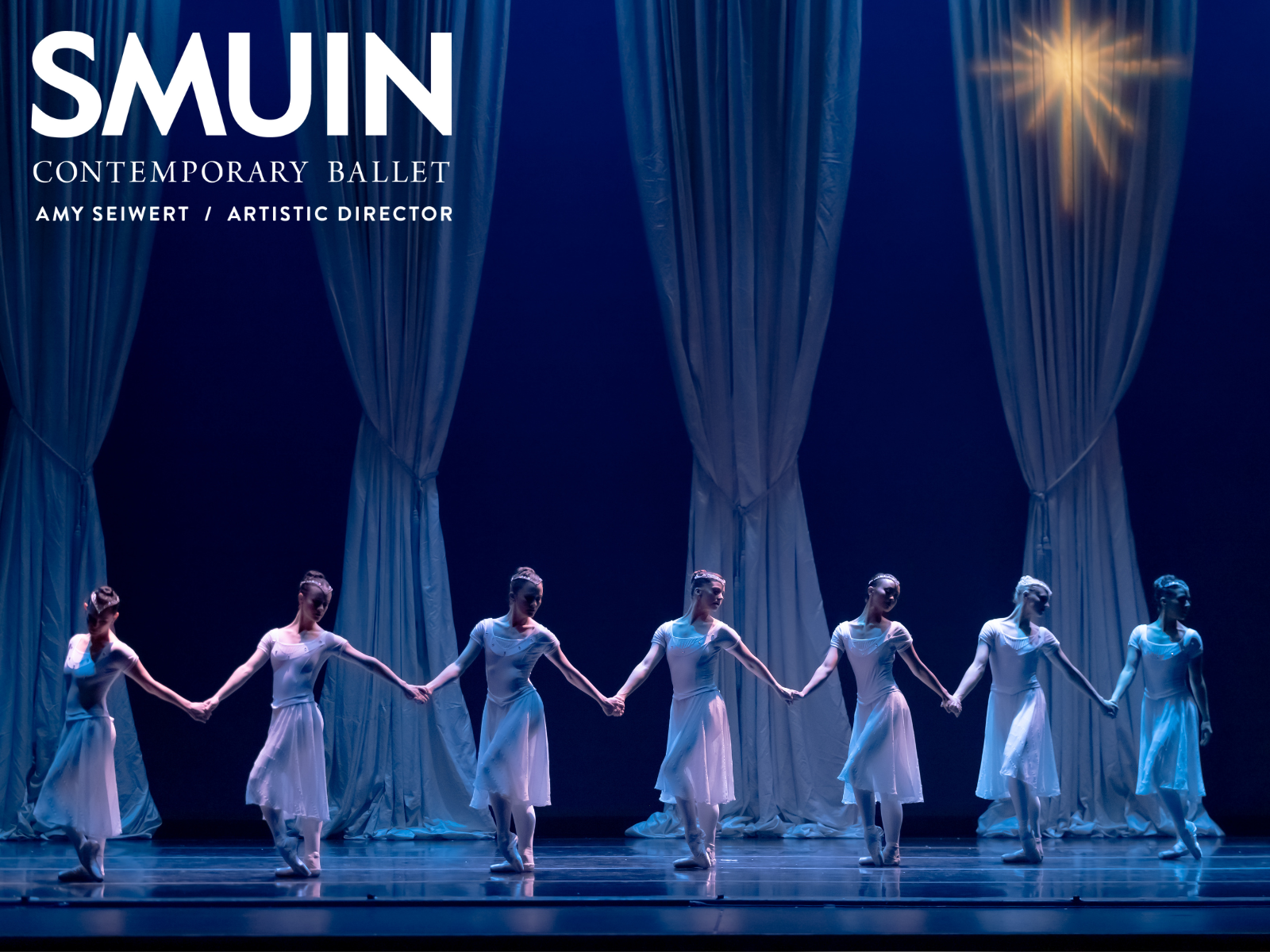 Seven ballet dancers in white costumes stand on stage holding hands in front of flowing white curtains under a star-like light. Text on the image reads "SMUIN Contemporary Ballet, Amy Seiwert / Artistic Director.