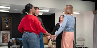 Four women stand in a circle holding hands inside a room with fluorescent lighting, chairs, and framed artwork in the background.