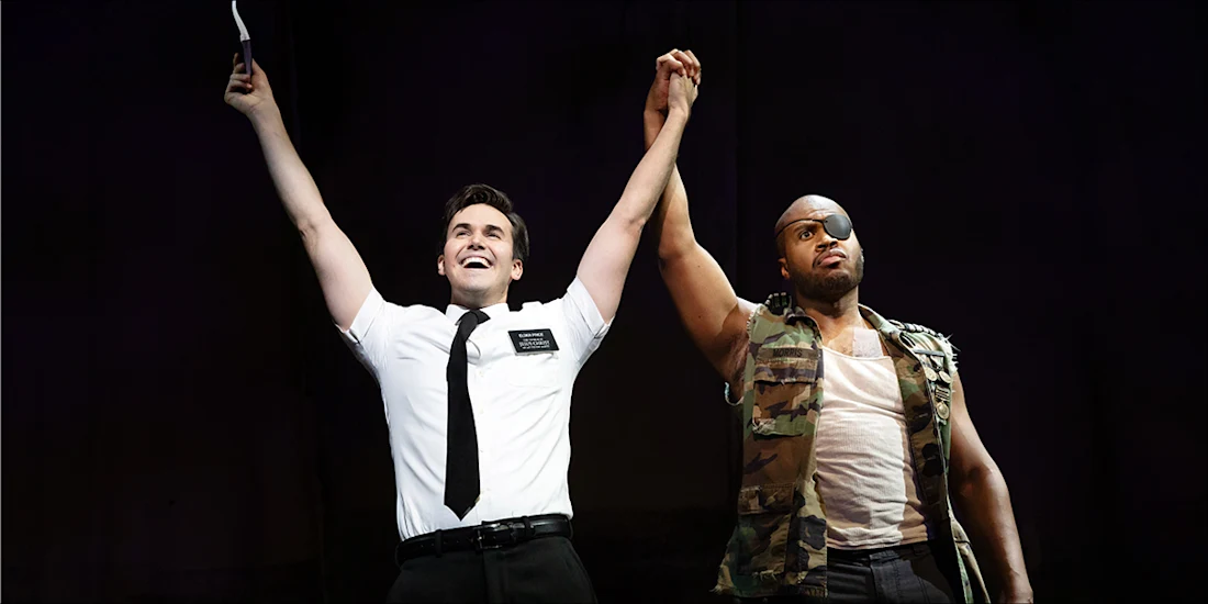 Two actors stand on stage holding hands; one wears a white shirt and tie, the other wears a camouflage vest and eyepatch.