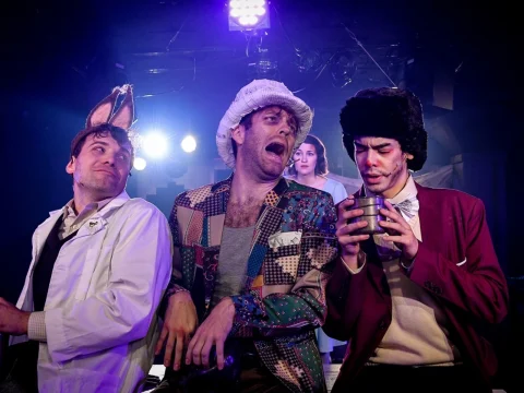 Three performers are on stage in costume: one in a lab coat, one in a colorful patchwork jacket and hat, and one in a suit with a fur hat. They are engaged in an energetic and expressive scene.