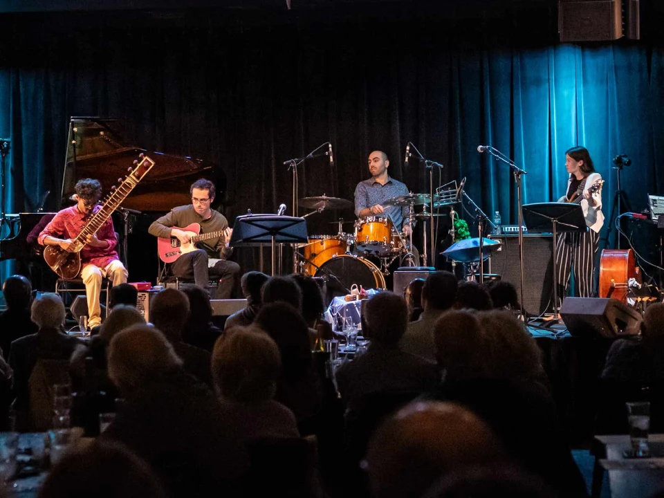 A band of five musicians performs on stage with various instruments including a sitar, guitar, double bass, drums, and violin. The audience watches attentively.