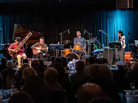 A band of five musicians performs on stage with various instruments including a sitar, guitar, double bass, drums, and violin. The audience watches attentively.