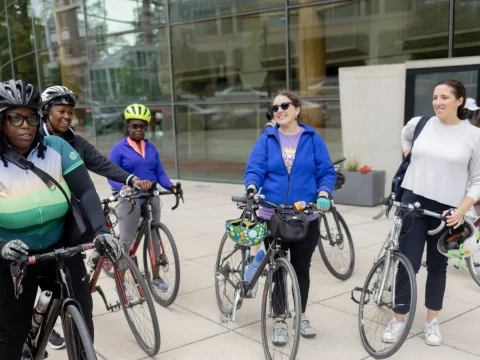 A group of women with bicycles stands outside a glass building. Some wear helmets and cycling gear, while others are in casual clothing.