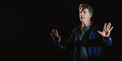 A man stands against a dark background, facing forward with both hands raised, wearing a blue jacket over a gray shirt and looking slightly to the side.