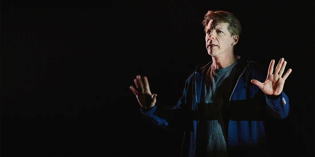 A man stands against a dark background, facing forward with both hands raised, wearing a blue jacket over a gray shirt and looking slightly to the side.
