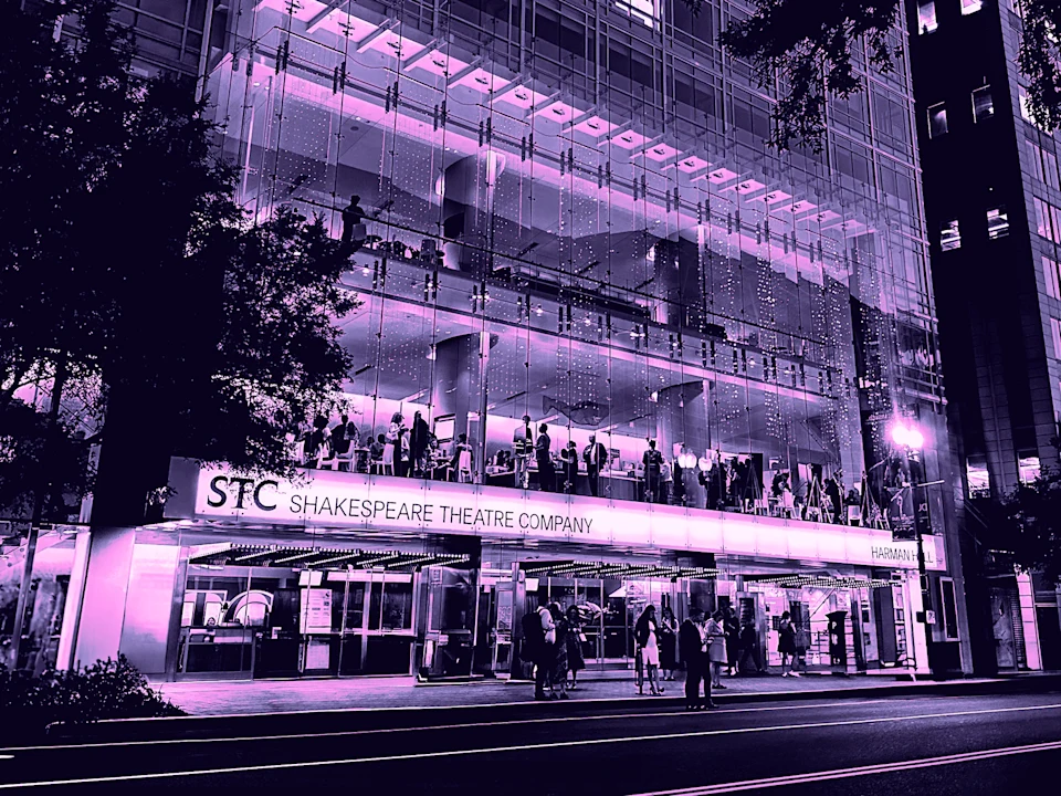 People gather outside and inside the glass-fronted building of the Shakespeare Theatre Company at night.