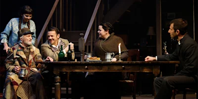 Five people in period clothing sit and stand around a wooden table with drinks, food, and candles, in a warmly lit room with a staircase in the background.