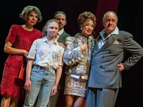 Five people stand together on stage in theatrical costumes, including a woman in a red dress, a girl in a white shirt and jeans, and others in formal and fashionable attire.