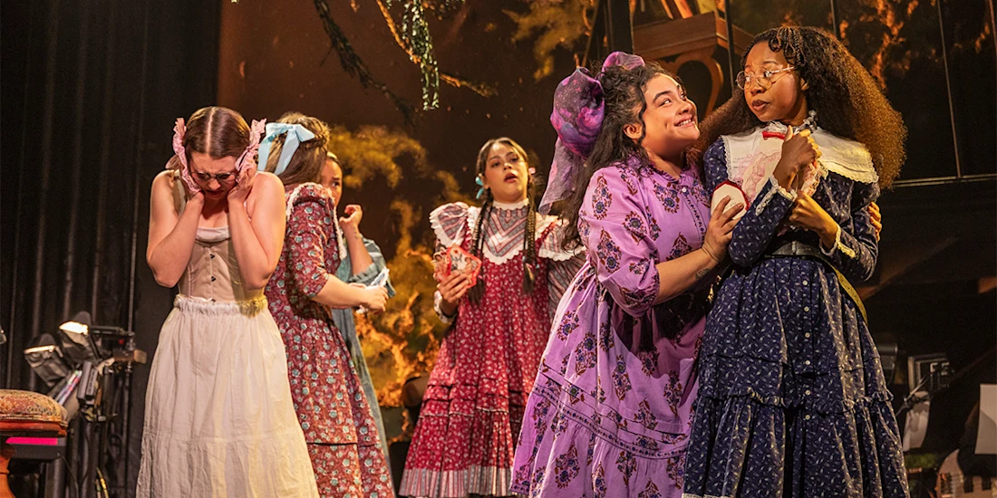 Six actors in period costumes perform on stage; some appear excited or affectionate, while one stands apart looking distressed, against a dramatic backdrop.