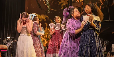 Six actors in period costumes perform on stage; some appear excited or affectionate, while one stands apart looking distressed, against a dramatic backdrop.