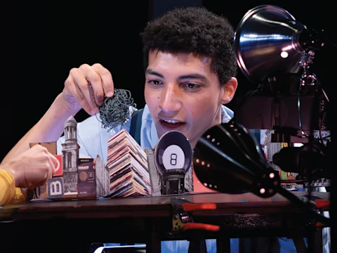 A person interacts with a table-top miniature city scene, holding a small wire object. Bright lights illuminate the setup.