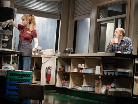 A woman in a maroon shirt works in a vintage diner kitchen, while a man in a plaid shirt sits at the counter drinking coffee.