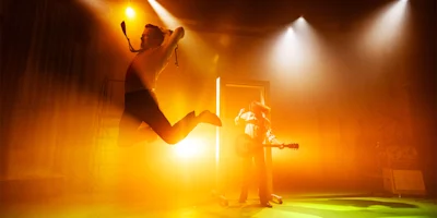Two people on a stage with dramatic lighting; one person jumps mid-air while the other stands with a guitar near an open door with bright light.