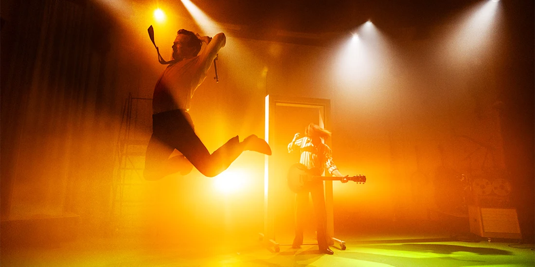Two people on a stage with dramatic lighting; one person jumps mid-air while the other stands with a guitar near an open door with bright light.