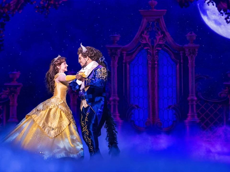 Two actors dressed as Belle and the Beast dance on stage, with a moonlit castle backdrop and blue lighting, recreating a scene from "Beauty and the Beast.