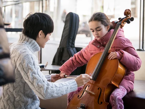 An adult helps a child play the cello inside a room. The child wears a pink jacket and patterned pants.