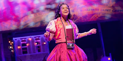 A young woman in a bright pink dress and jacket stands on stage, smiling, with a camera around her neck; colorful stage lights and a backdrop are visible behind her.
