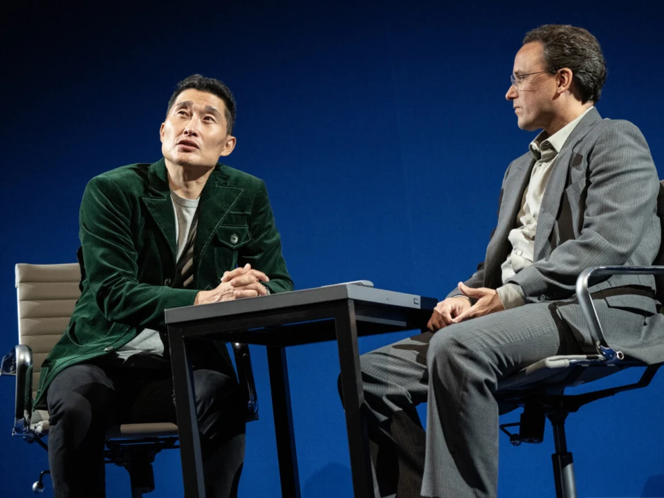 Two men sit at a table on stage against a blue background. One wears a green jacket and the other a gray suit.