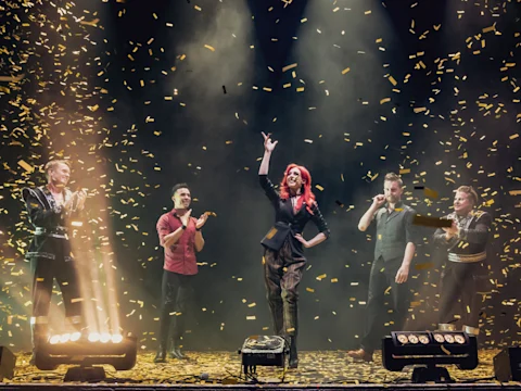 Performers stand on a stage surrounded by falling confetti, with one person at center stage raising a hand triumphantly under spotlights.