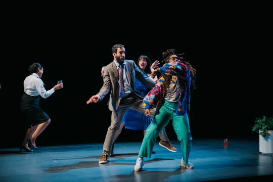 A group of actors performs energetically on stage, with two actors in the foreground appearing to fend off an unseen threat, while others in the background look engaged in the scene.