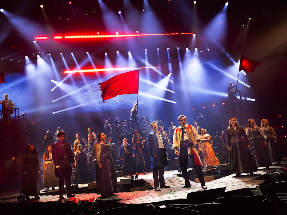 A large theater cast performs on stage with dramatic lighting; one actor holds a red flag while others stand in period costumes, evoking a revolution scene.