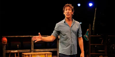 A man in a blue shirt stands on stage, gesturing with one hand, with stage props and dim lighting in the background.