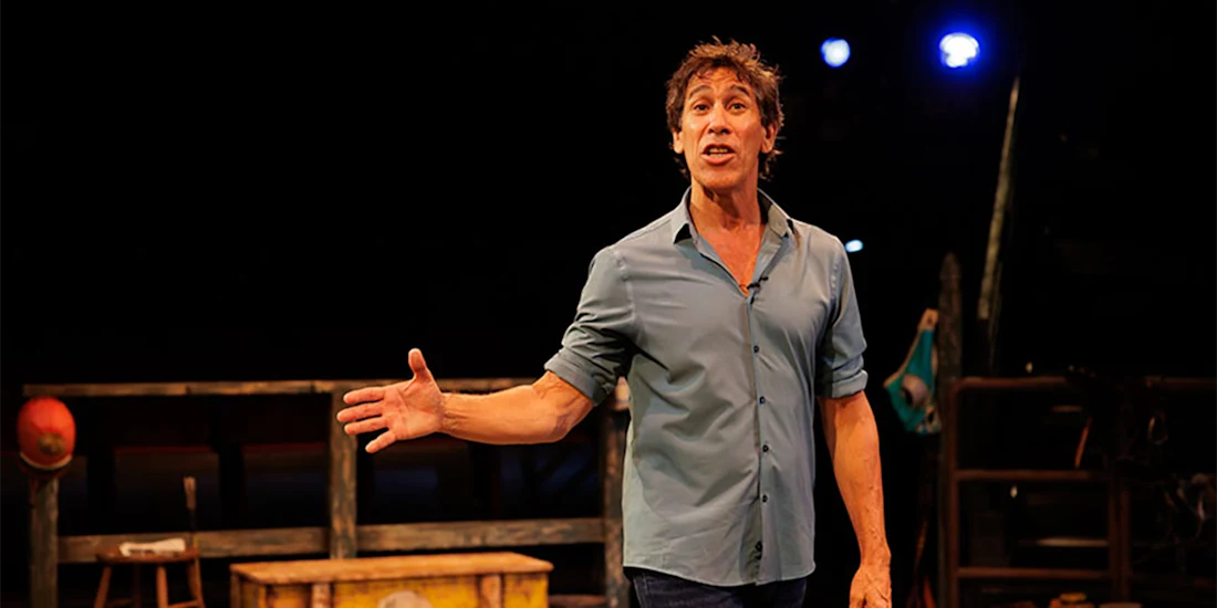 A man in a blue shirt stands on stage, gesturing with one hand, with stage props and dim lighting in the background.