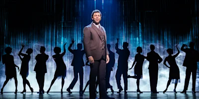 A man in a suit stands center stage, spotlighted, with a group of people silhouetted in dance poses behind him against a blue-lit backdrop.