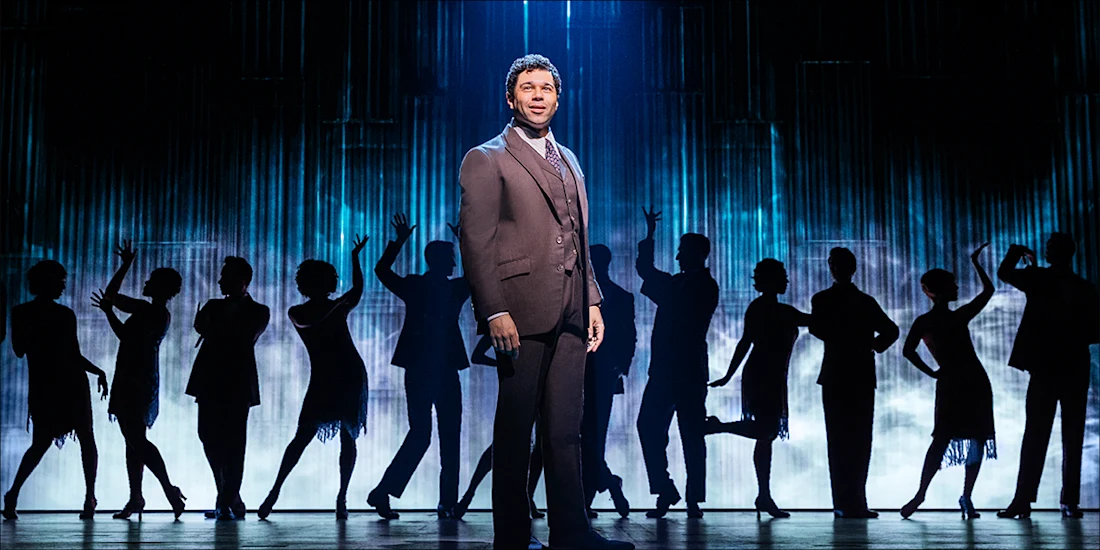 A man in a suit stands center stage, spotlighted, with a group of people silhouetted in dance poses behind him against a blue-lit backdrop.