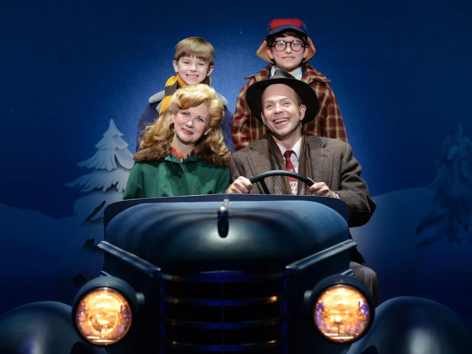 A family of four dressed in vintage winter clothing sit in a car on a stage set with a painted winter backdrop.