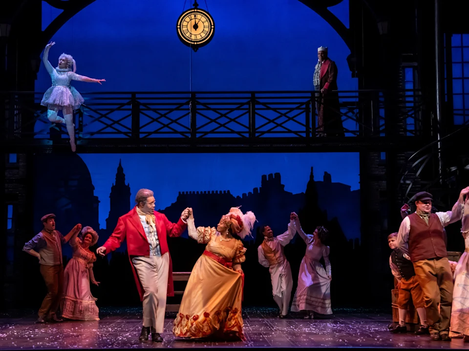 Actors perform a lively scene on stage in period costumes, with a clock overhead and blue-lit cityscape backdrop; a woman in white is suspended above the ensemble.