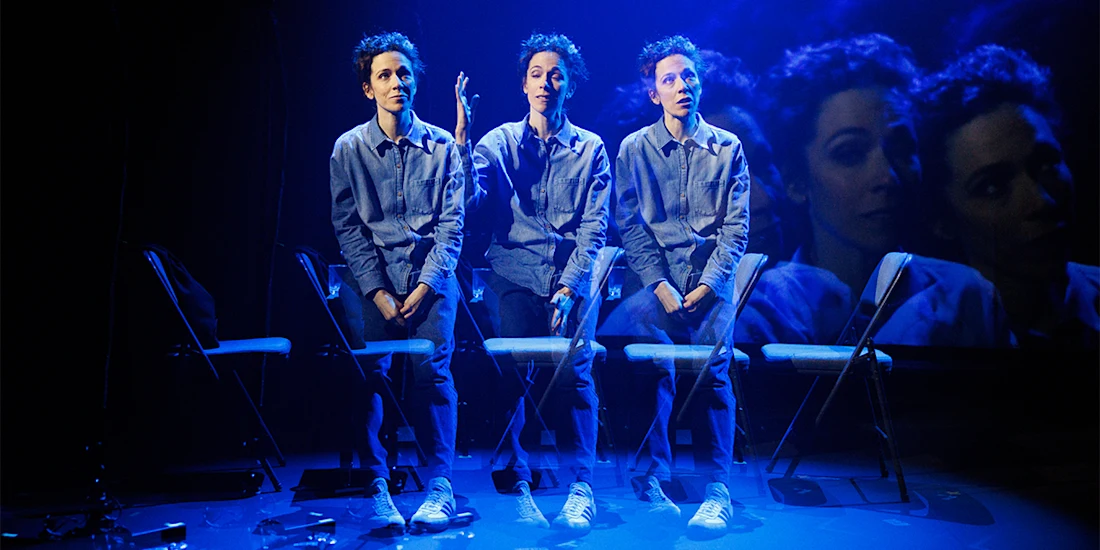 A person in a blue shirt and pants is shown sitting in three overlapping positions on chairs, with a large projected face in the background, under blue stage lighting.
