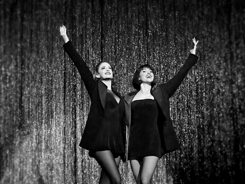 Two women in black outfits stand side by side on stage with their arms raised, smiling, in front of a shimmering curtain.